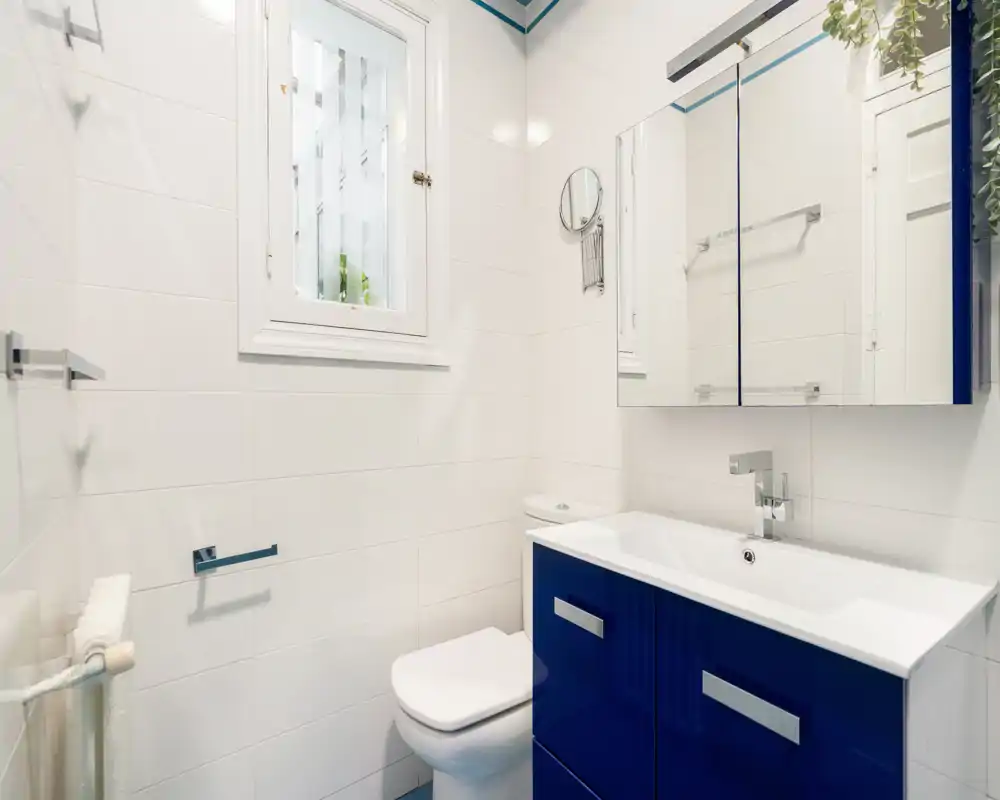 Baño moderno y luminoso con paredes alicatadas blancas, inodoro compacto y mueble de lavabo azul brillante con espejo encima.