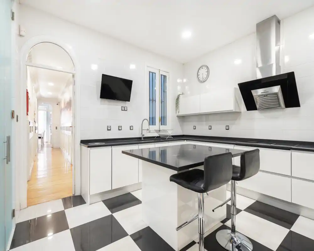 Cocina moderna y luminosa con isla central, taburetes y acabados elegantes en blanco y negro. Espacio limpio y bien iluminado, ideal para cocinar y comer informalmente.