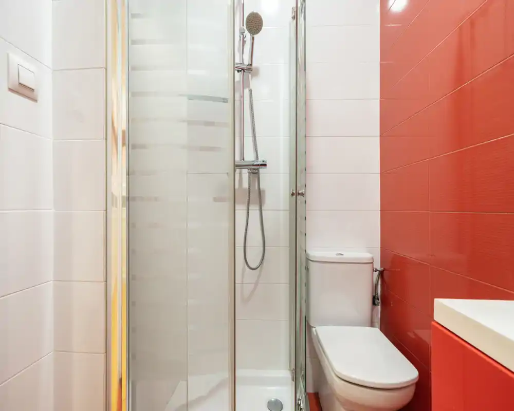 Baño moderno y compacto con ducha acristalada, cabezal de ducha mural, inodoro y azulejos rojos y blancos que ofrecen un aspecto contemporáneo y luminoso.
