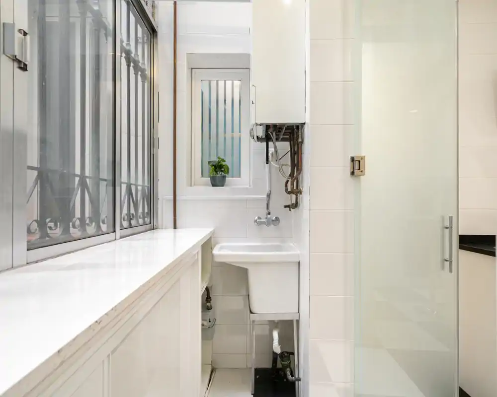 Rincón de utilidad compacto con un fregadero mural y un calentador junto a una gran ventana. Azulejos y encimera blancos que aportan un espacio luminoso y funcional, ideal para labores de lavado o servicio.