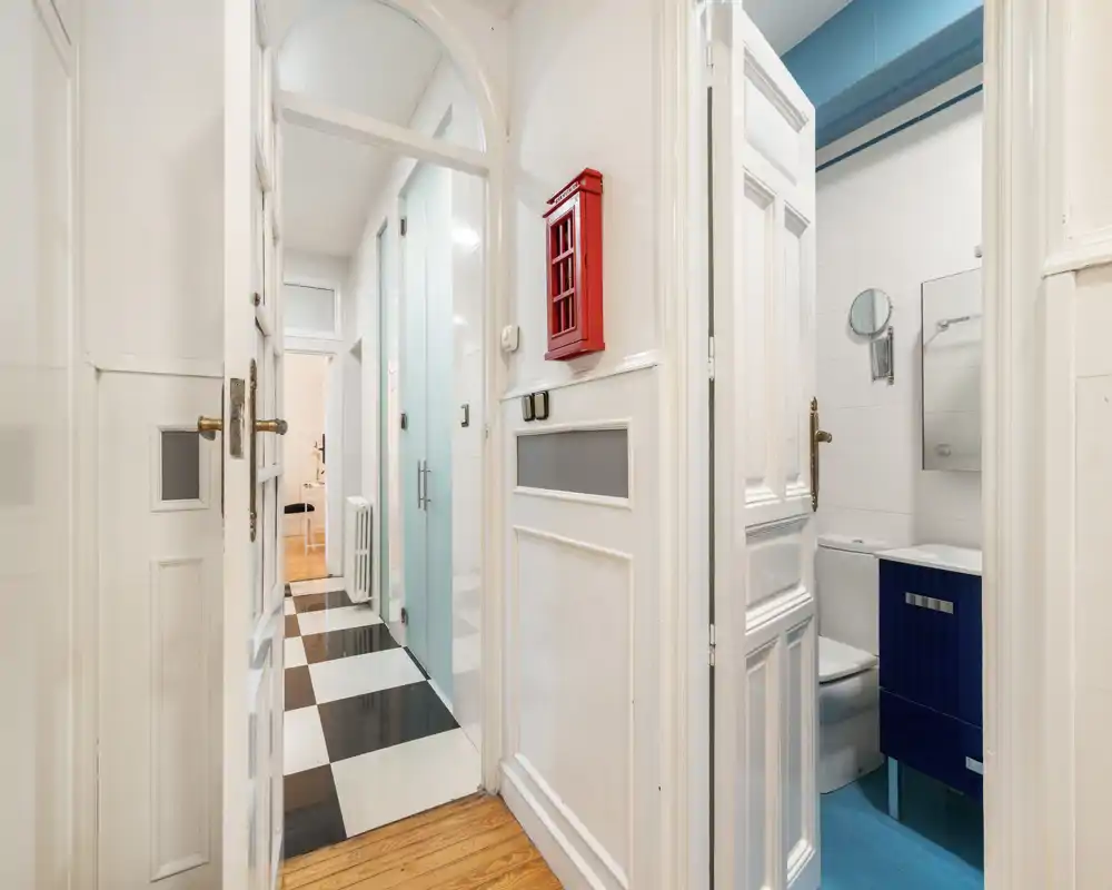 Entrada/pasillo luminoso y estrecho con puertas blancas decorativas, suelo ajedrezado en blanco y negro y un pequeño baño visible a la derecha.