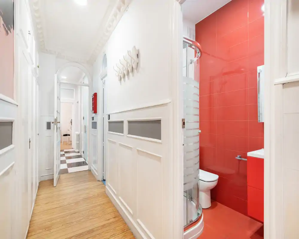 Entrada/pasillo luminoso con panelado blanco decorativo, suelo de madera y puertas que dan a habitaciones adyacentes, incluido un baño con azulejos rojos.