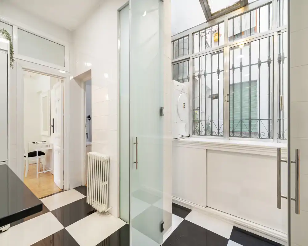 Cocina/área de lavandería luminosa y funcional con ventana grande, suelo ajedrezado, radiador y lavadora-secadora apiladas; espacio limpio y práctico para el uso diario.