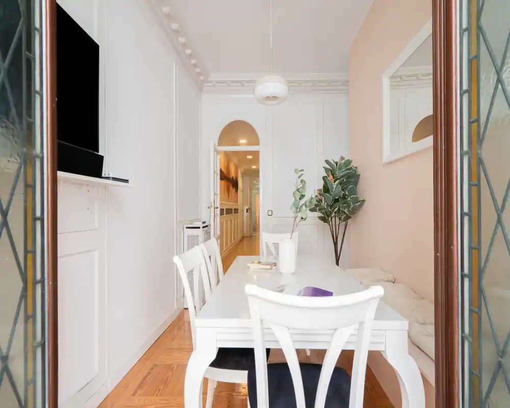 Zona de comedor luminosa y elegante con mesa y sillas blancas, banco, lámpara colgante y planta interior — ideal para comidas relajadas en un apartamento con estilo.