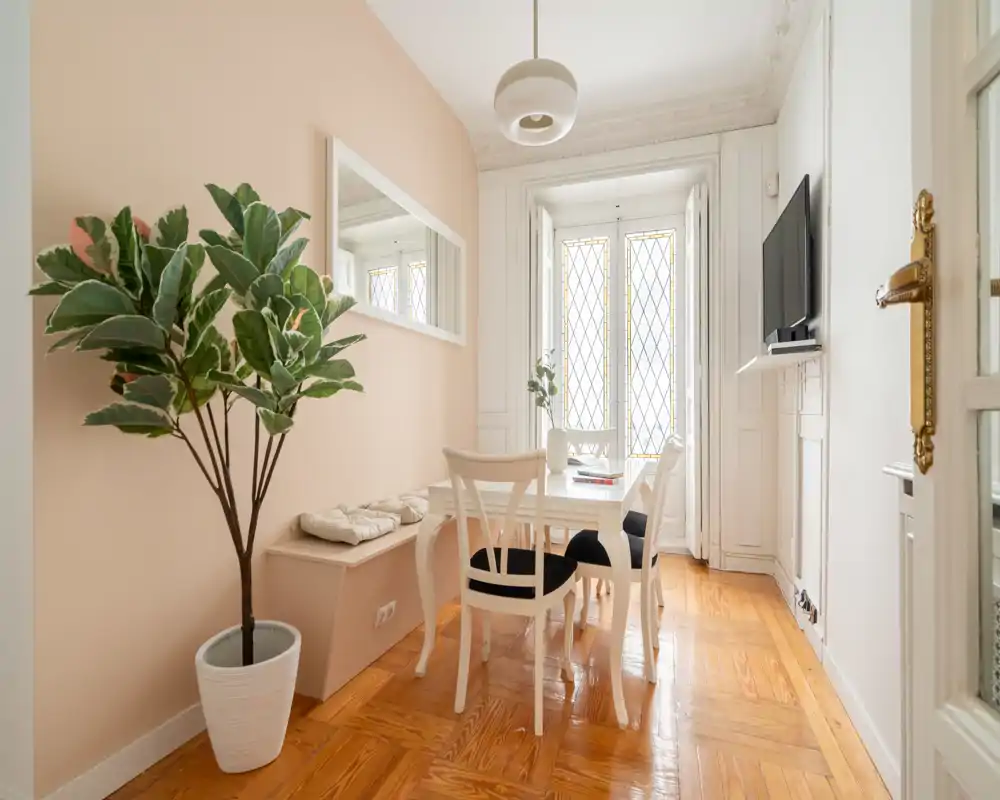 Acogedor comedor luminoso con mesa y sillas blancas, banco decorativo y una gran planta en maceta; suelo de madera cálida y ventanas altas que aportan mucha luz natural.