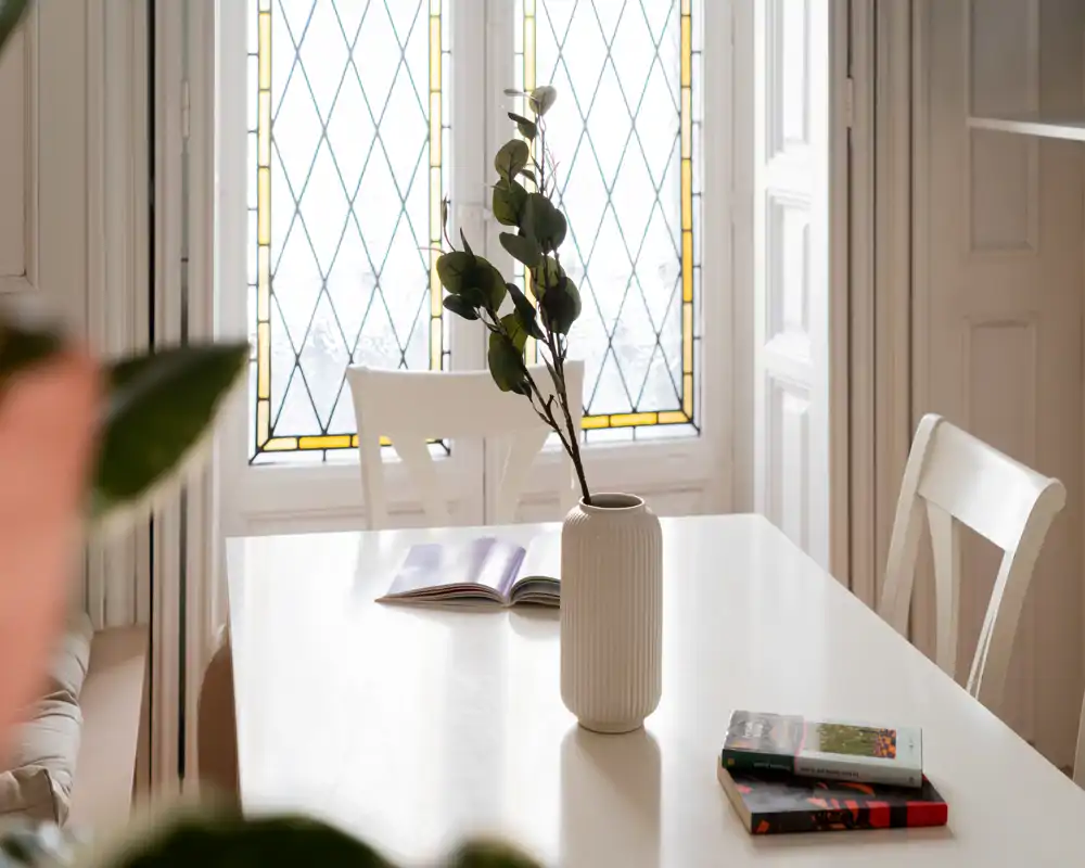 Zona de comedor luminosa y estilosa con mesa blanca, sillas y un jarrón decorativo frente a una ventana con vidrio decorado — ideal para desayunos tranquilos o lectura.