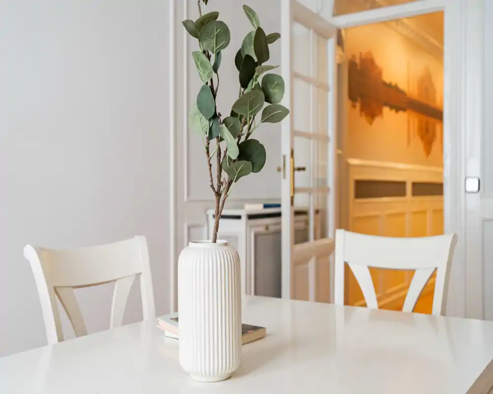 Zona de comedor luminosa y minimalista con mesa y sillas blancas y un jarrón decorativo — estilo limpio y moderno, ideal para anuncios de pisos.