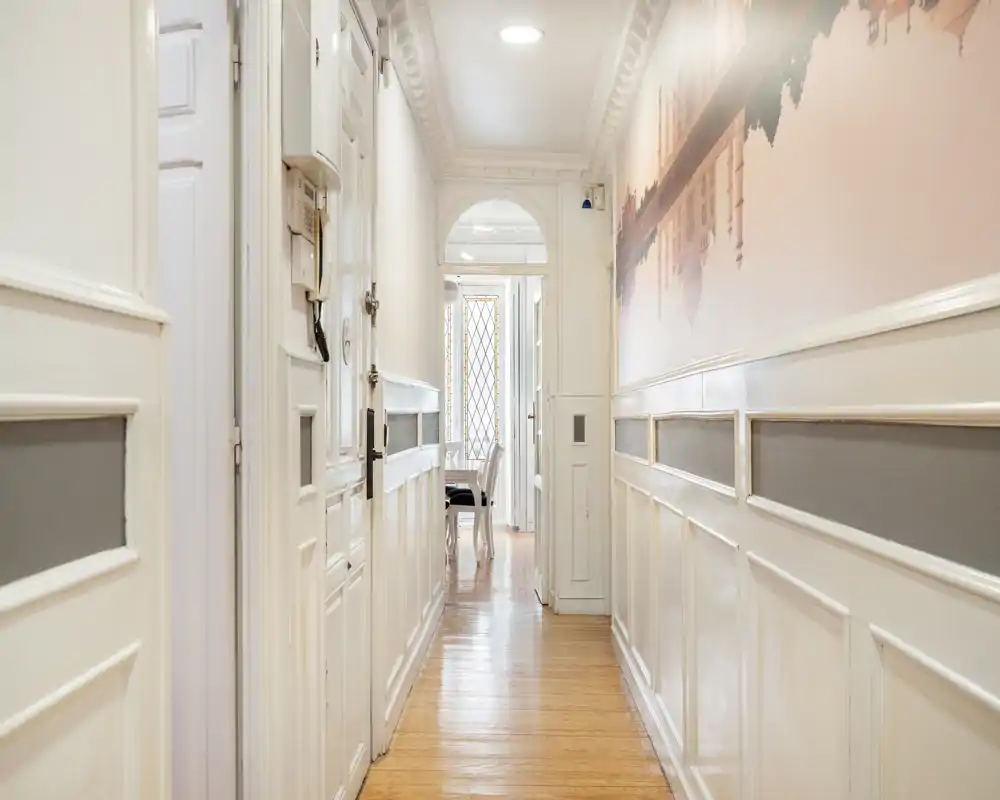 Vestíbulo/pasillo luminoso y estrecho con paneles blancos, molduras decorativas y suelo de madera, que conduce a una habitación iluminada.