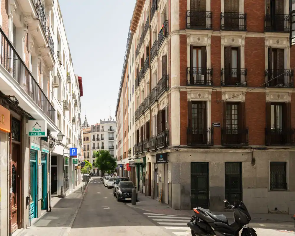 Escena de calle urbana soleada con edificios residenciales de varios pisos, coches aparcados y una moto en primer plano; ideal para mostrar el barrio y los exteriores.