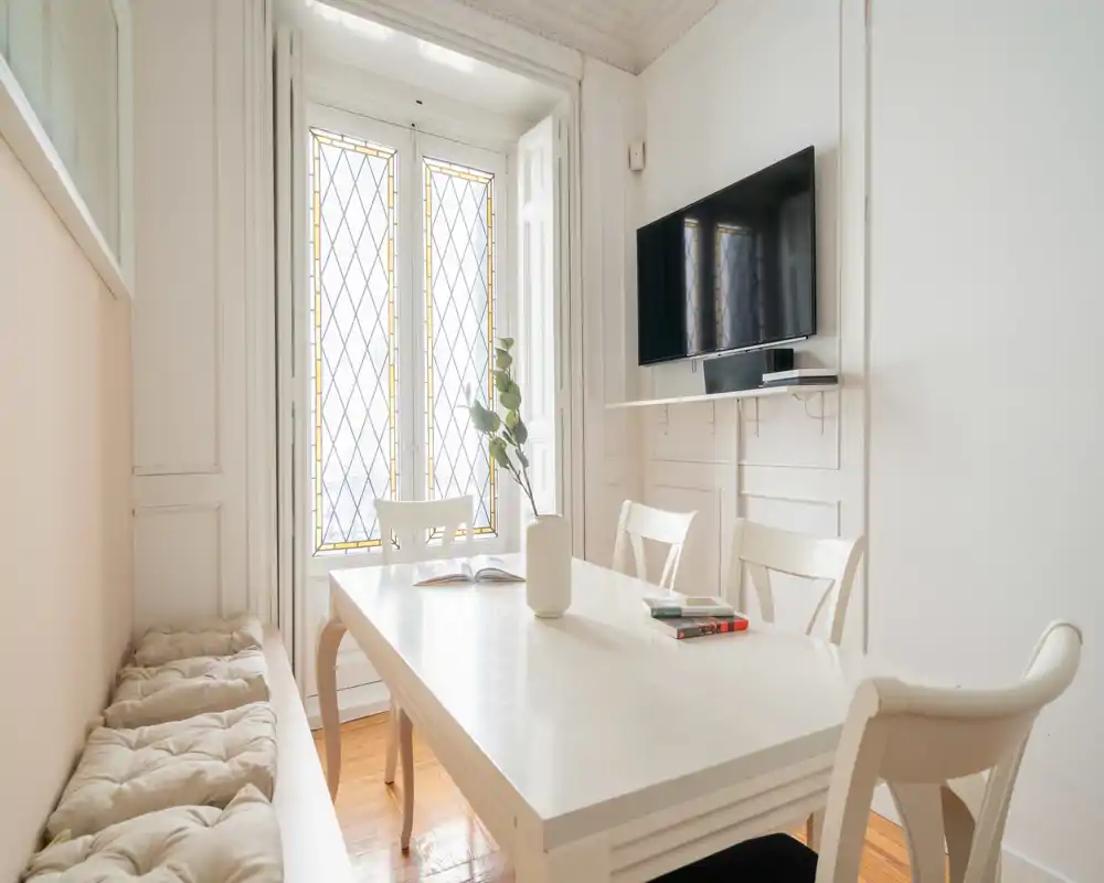 Zona de comedor luminosa y elegante con mesa blanca, banqueta acolchada y sillas junto a altas ventanas con cristales decorativos; decoración minimal y luz natural que crean un ambiente acogedor.