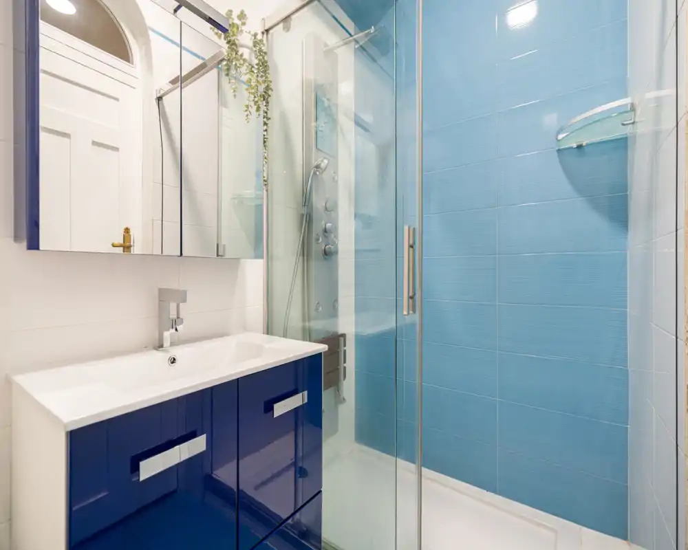 Baño moderno y luminoso con ducha acristalada, pared con azulejos azules y mueble de lavabo lacado azul marino con espejo-armario.