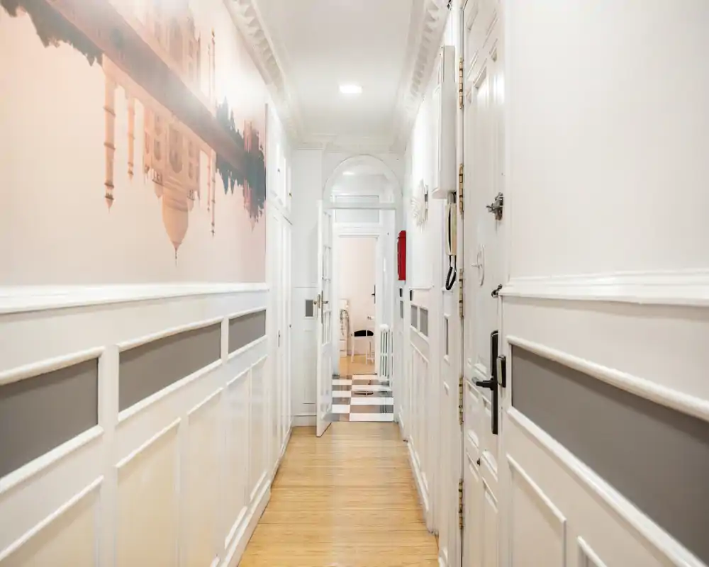 Entrada luminosa y estrecha con paredes blancas paneladas, un mural decorativo y suelo de madera que conduce a las habitaciones interiores.