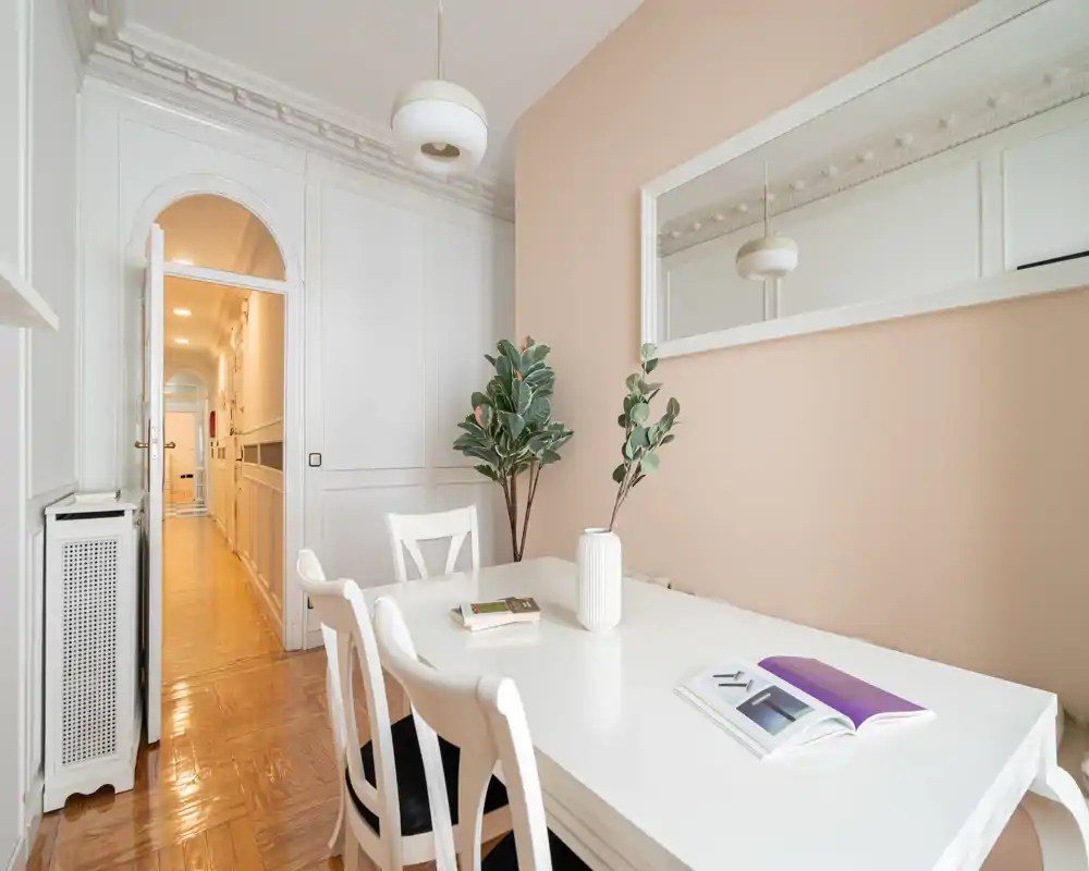 Comedor luminoso y elegante con mesa y sillas blancas, pared color melocotón, plantas decorativas y un gran espejo que crea un ambiente acogedor.