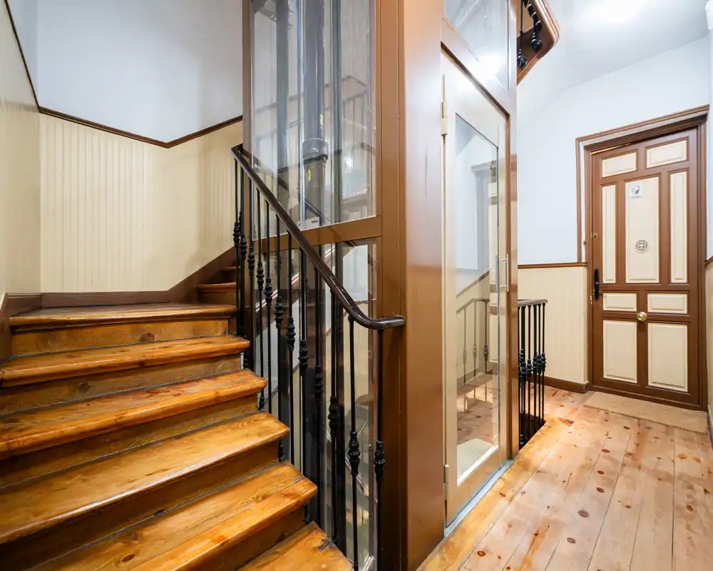 Entrada bien iluminada con escaleras de madera pulida, un pequeño hueco de ascensor y una puerta decorativa con paneles — suelos de madera cálida y luz nítida dan una sensación acogedora.