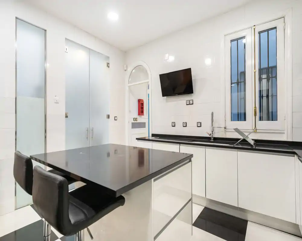 Cocina moderna y luminosa con isla central y dos taburetes, encimeras negras y armarios blancos que ofrecen un aspecto limpio y contemporáneo.
