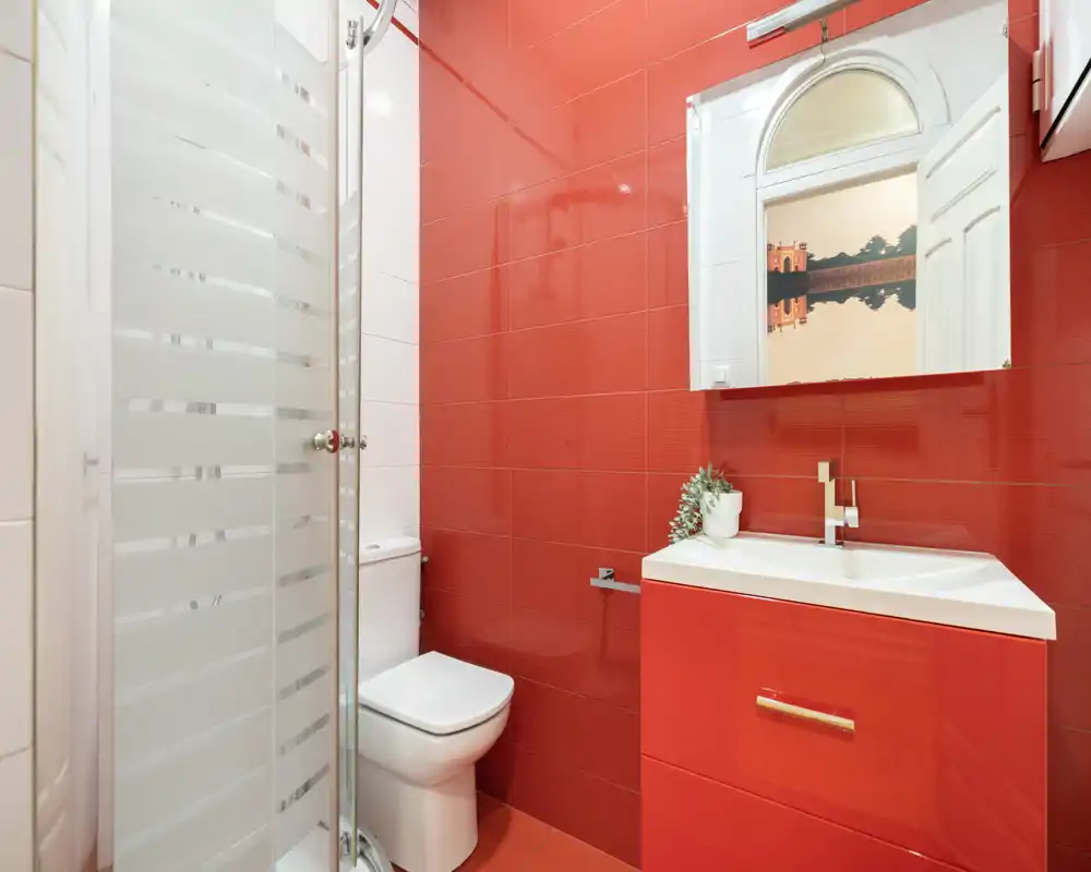 Baño moderno y compacto con azulejos rojos brillantes, ducha de esquina con mampara de cristal, inodoro y lavabo con mueble blanco. Espacio limpio, elegante y bien iluminado, ideal para alquileres de corta estancia.