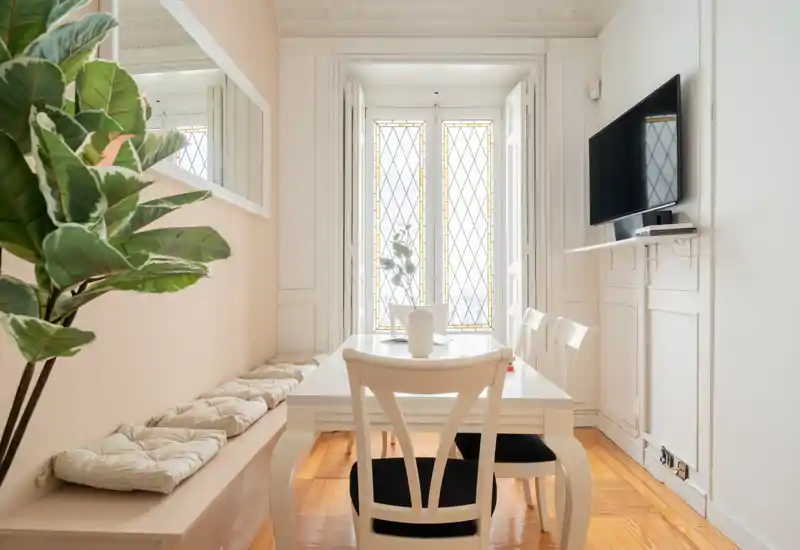 Accogliente angolo pranzo luminoso con tavolo bianco, panca con cuscini e una finestra con vetro colorato decorativo. Pavimenti in legno caldo e una grande pianta rendono l'ambiente invitante.