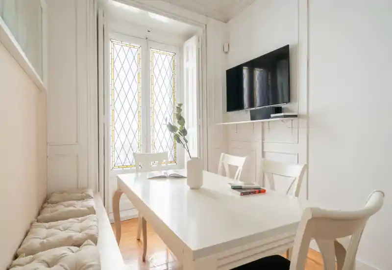 Coin repas lumineux et élégant avec table blanche, chaises et banquette près d'une grande fenêtre ; quelques touches décoratives (vase, livres) pour une ambiance calme et accueillante.