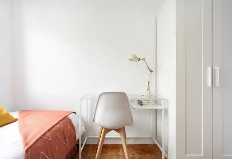 Coin chambre lumineux et minimaliste avec lit, couverture chaude et petit bureau avec lampe — idéal pour un séjour confortable ou le télétravail.