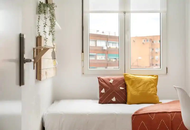 Chambre lumineuse au style épuré avec un lit simple sous une grande fenêtre, coussins chaleureux et plaid créant une atmosphère accueillante.