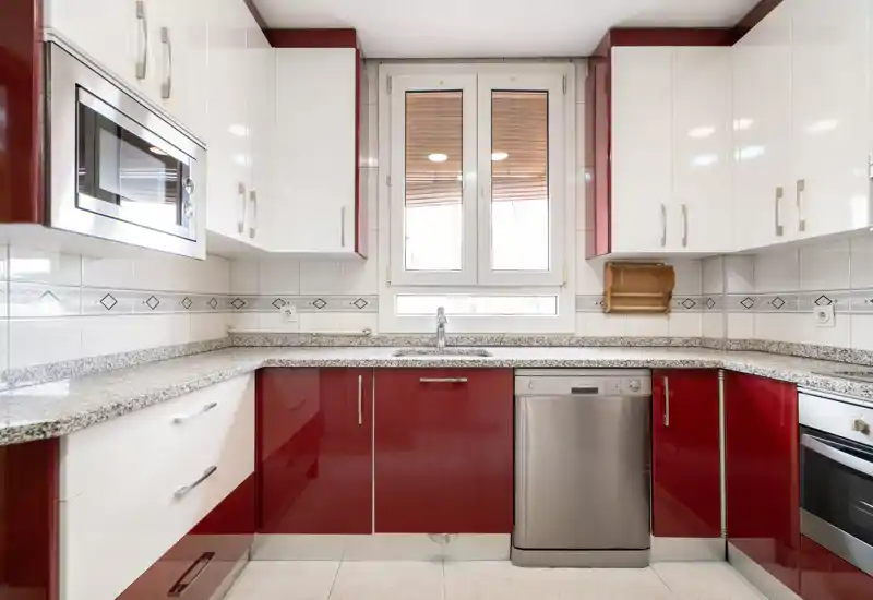 Cuisine moderne et lumineuse avec meubles rouges et blancs, plans en granit, micro-ondes et lave-vaisselle intégrés, et une fenêtre centrale laissant entrer la lumière naturelle.