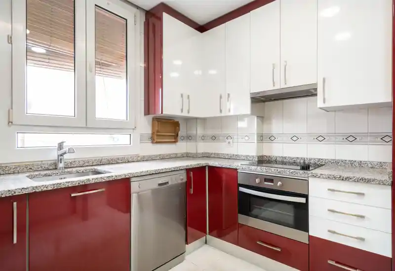 Cuisine contemporaine avec meubles rouges et blancs, plans de travail imitation granit, four et lave-vaisselle encastrés, et une fenêtre lumineuse apportant de la lumière naturelle.