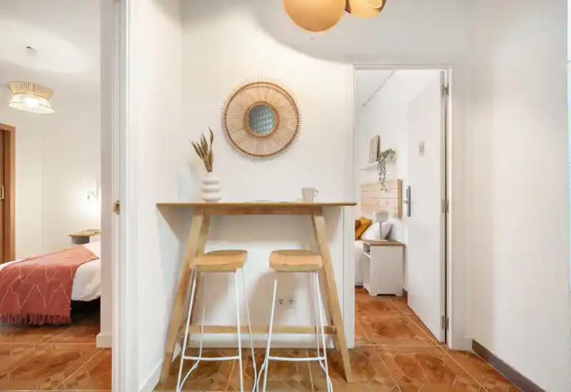 Scène de chambre lumineuse et chaleureuse avec un petit bar petit-déjeuner au premier plan. On distingue partiellement deux lits à travers les portes, des textiles doux et des accents en bois naturel, pour une ambiance confortable adaptée aux annonces locatives.
