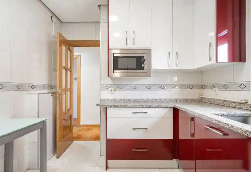 Cuisine moderne et lumineuse avec placards laqués rouge et blanc, plans de travail en granit et micro-ondes encastré. Carrelage propre et agencement compact, parfait pour une cuisine fonctionnelle.