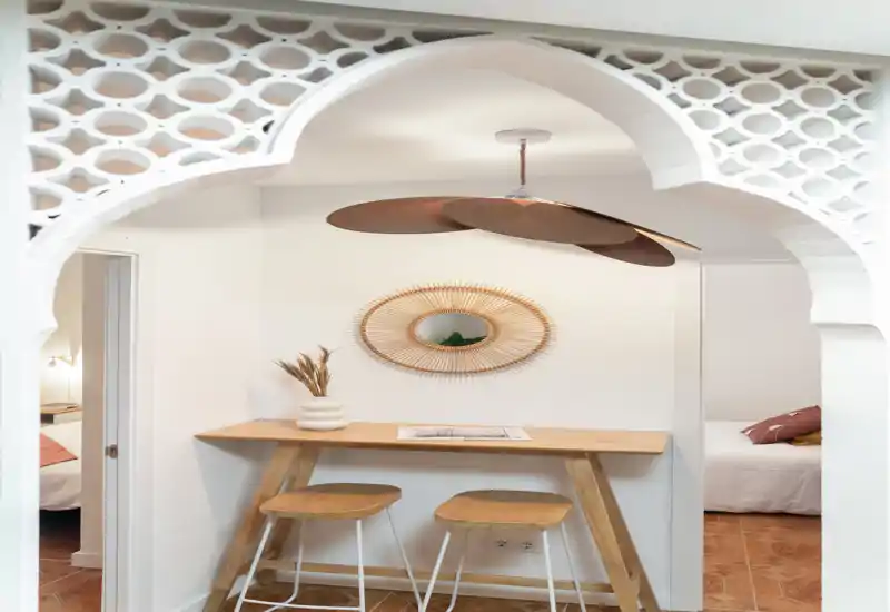 Niche ouverte et élégante avec un bar en bois et deux tabourets encadrée par un arc ornemental. La décoration chaleureuse, le miroir en rotin et la lumière douce créent un espace de vie accueillant, parfait pour un repas rapide ou du télétravail.