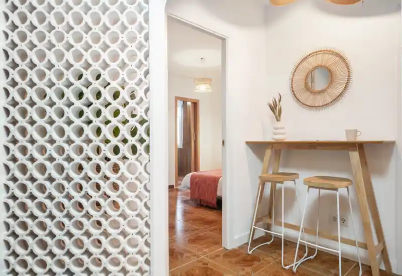 Chambre accueillante et lumineuse visible par une porte, lit avec un plaid chaleureux ; au premier plan, un comptoir avec tabourets et un miroir décoratif pour une ambiance moderne.