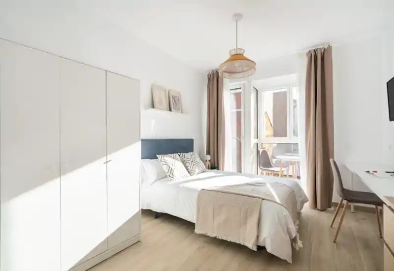 Bright, modern bedroom with a double bed, soft textiles and natural light pouring from a balcony window. Neutral tones and minimalist decor create a calm, inviting atmosphere.