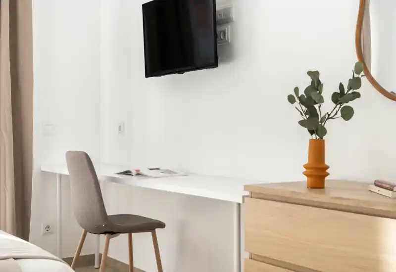 Bright, minimal bedroom corner with a wall-mounted TV, compact desk and a chair. Soft neutral tones, wooden dresser and a decorative vase create a calm, modern feel ideal for rental photos.