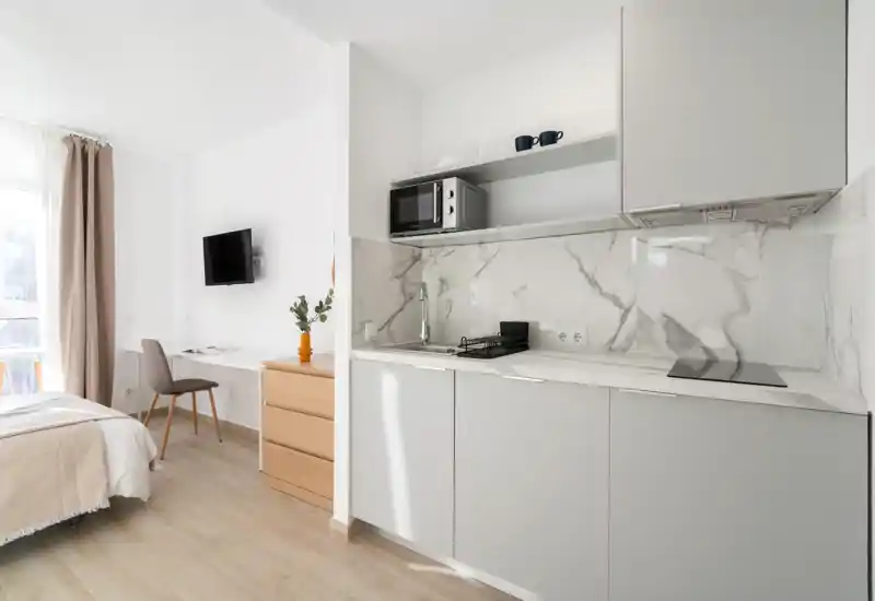 Sunny modern studio bedroom with an integrated kitchenette. Calm neutral tones, a compact cooking area with marble-effect backsplash, a comfy bed near a bright window and a small work corner.