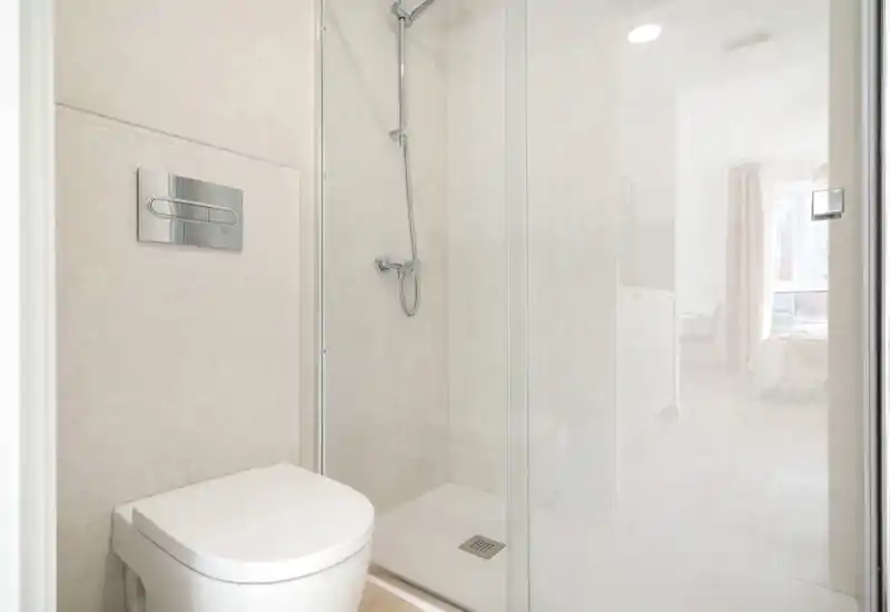 Bright, minimalist bathroom with a wall-mounted flush plate, compact toilet and glass-enclosed walk-in shower — clean neutral tiles and natural light from an adjacent room.