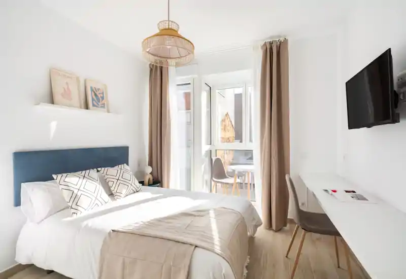 Bright, well-styled bedroom with a comfortable double bed, soft neutral linens, a desk workspace and lots of natural light from the balcony door.