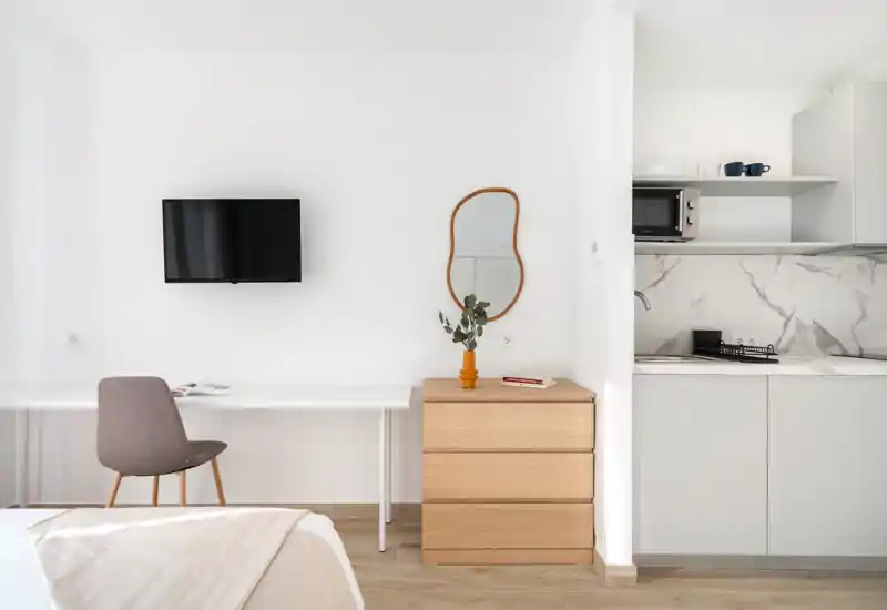 Bright, minimalist bedroom corner with a visible bed corner, desk, wall-mounted TV and a compact kitchenette. Clean lines, neutral tones and simple wooden furniture give a calm, modern feel.