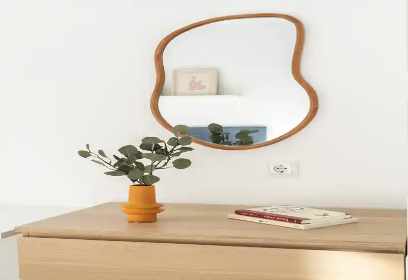 Minimal, well-lit console surface with a sculptural wooden-framed mirror and a small plant in an ochre pot — a calm, modern entry vignette.