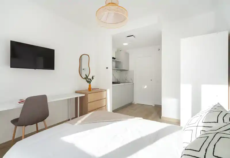 Bright, minimal bedroom with a cozy bed, wall-mounted TV, desk and a small kitchenette visible through an open alcove — ideal for a studio rental with modern, clean styling.