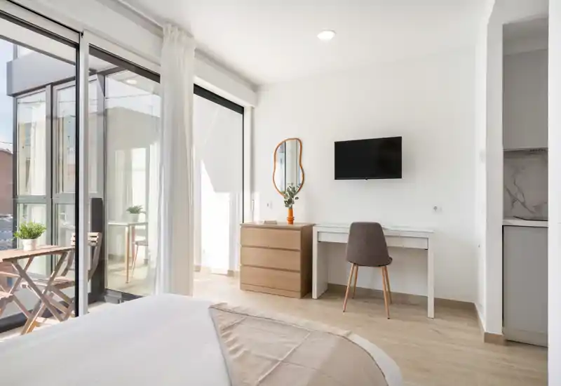 Bright, minimal bedroom with a comfortable bed in the foreground, a desk and dresser against the wall, and large sliding glass doors opening to a small balcony.