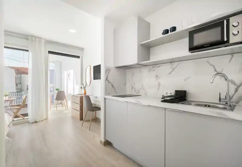 Modern kitchenette with marble-effect backsplash, integrated sink and induction hob; bright, minimal and well-lit space opening onto a sunlit balcony.