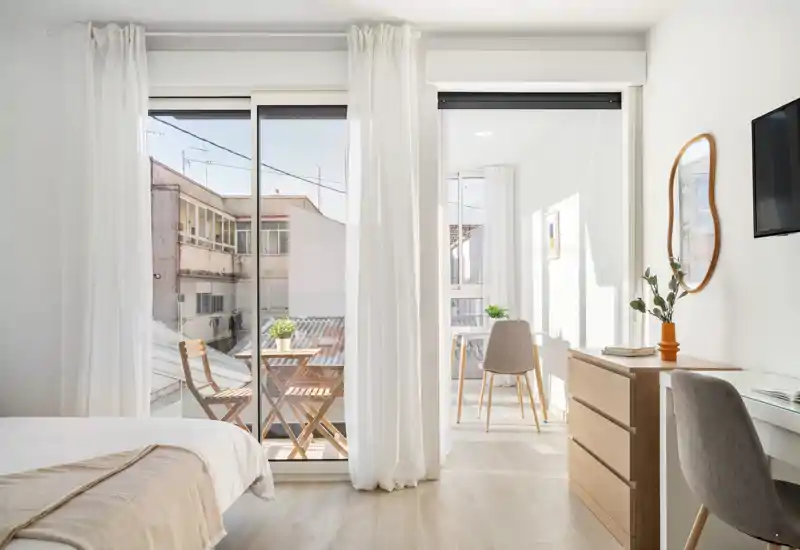 Bright, modern bedroom with a comfortable bed, light wood furniture, a small balcony with outdoor chairs and abundant natural light — ideal for restful stays.