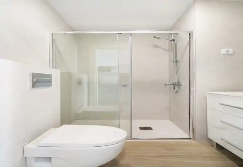 Bright modern bathroom with a glass-enclosed shower, wall-mounted toilet, and a sleek white vanity — clean minimalist design with wood-effect flooring.