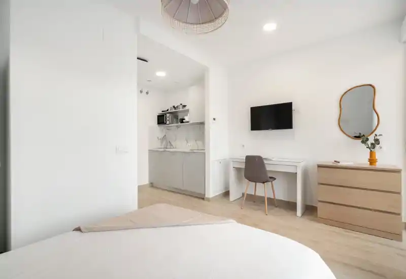 Bright, modern bedroom with a partial view of a neatly made bed in the foreground, a small kitchenette alcove, wall-mounted TV, desk and dresser — minimalist and well lit for short-stay rentals.