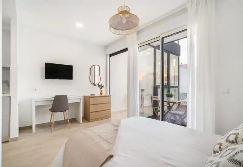 Bright, modern bedroom with a comfortable bed, a desk and dresser, and large sliding doors opening to a small furnished balcony.