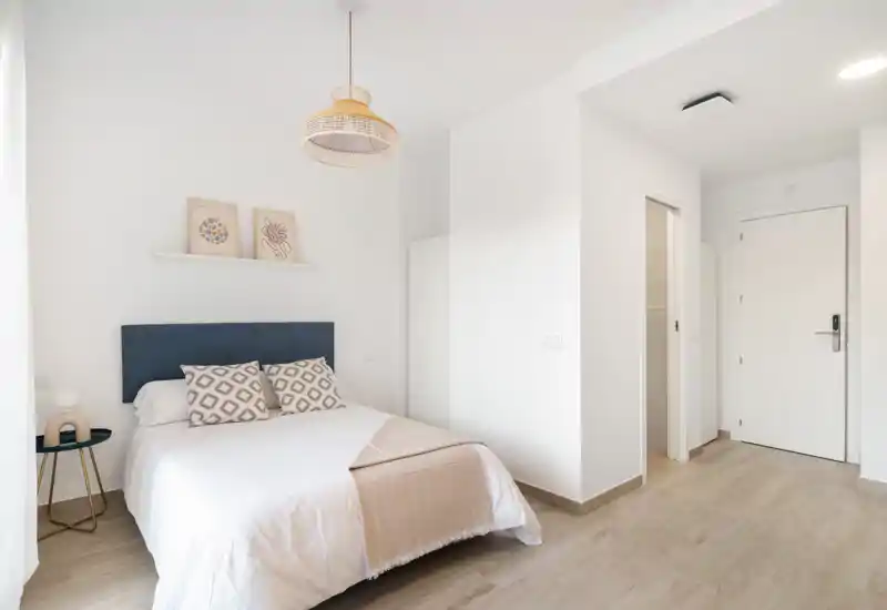 Bright, minimal bedroom with a double bed, patterned cushions, a blue headboard and soft natural light from the left. The neutral palette and simple decor create a calm, inviting atmosphere ideal for rental listings.