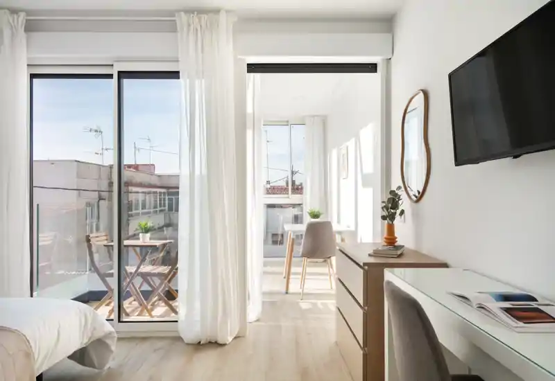 Chambre moderne et lumineuse avec un lit confortable au premier plan, lumière naturelle via une porte-fenêtre menant à un petit balcon, espace bureau et mobilier en bois élégant.