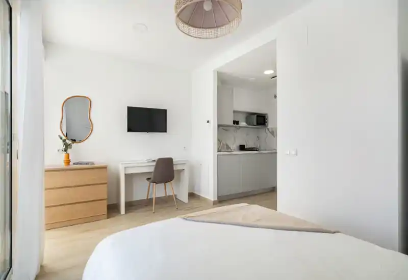 Chambre moderne et lumineuse avec lit blanc au premier plan, petit bureau avec TV murale et kitchenette compacte visible par une ouverture.