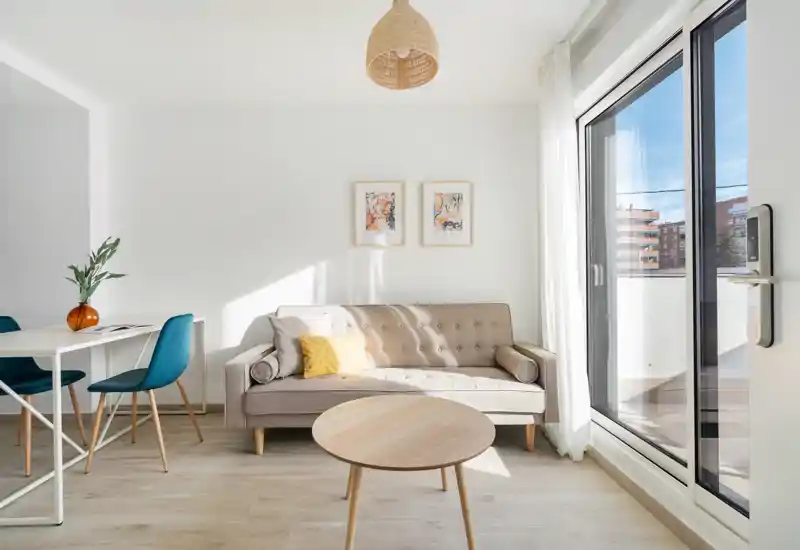 Bright, modern living area with a tufted sofa, round wooden coffee table and large sliding glass door that opens to a balcony—clean Scandinavian-style decor and plenty of natural light.