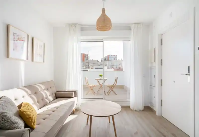 Bright, stylish living room with a light-colored tufted sofa, round wooden coffee table and sliding doors opening to a small terrace — airy, modern and inviting.