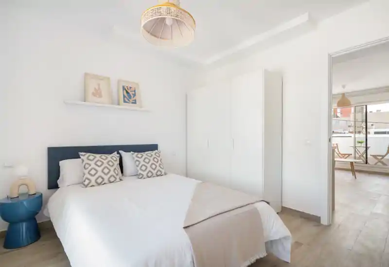 Bright, minimal bedroom with a double bed, decorative pillows, wardrobe and natural light flowing from an adjacent room — ideal for rental listings highlighting modern simplicity and comfort.
